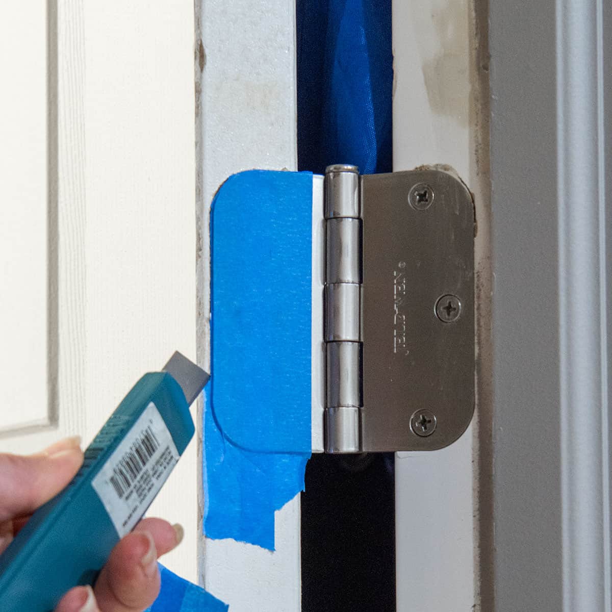 Painters tape around a door hinge being cut in preparation for painting.