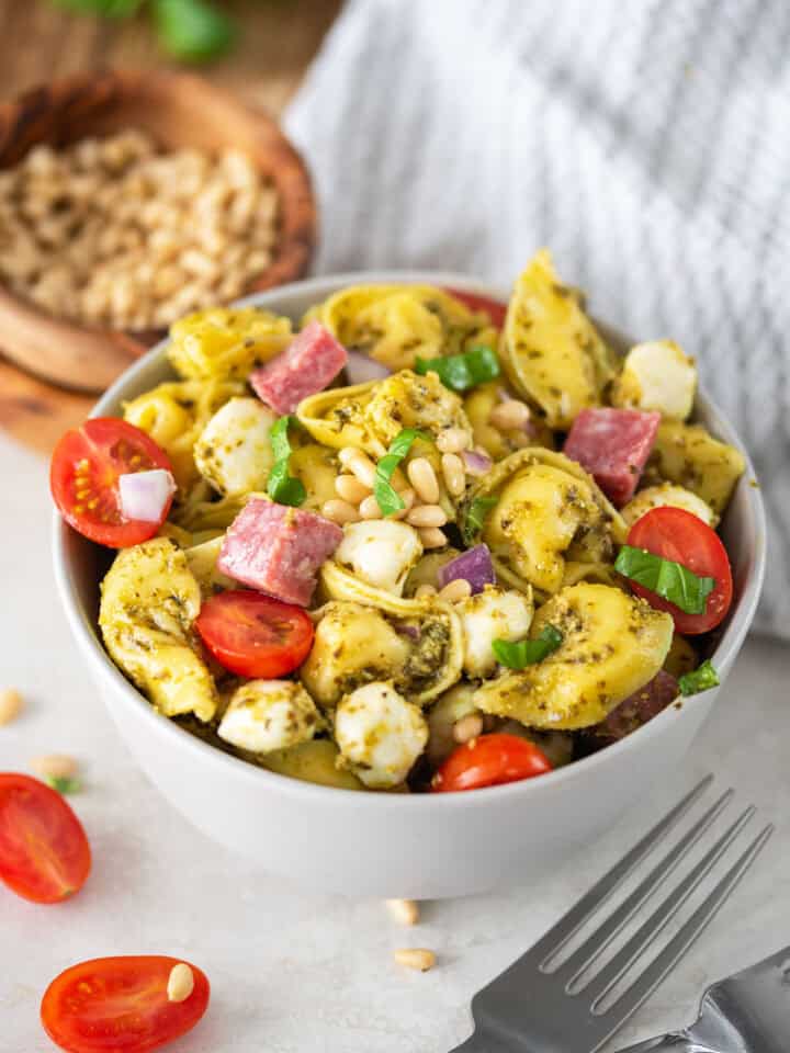 A bowl of pesto tortellini pasta served cold with mozzarella, salami, and tomatoes on top.